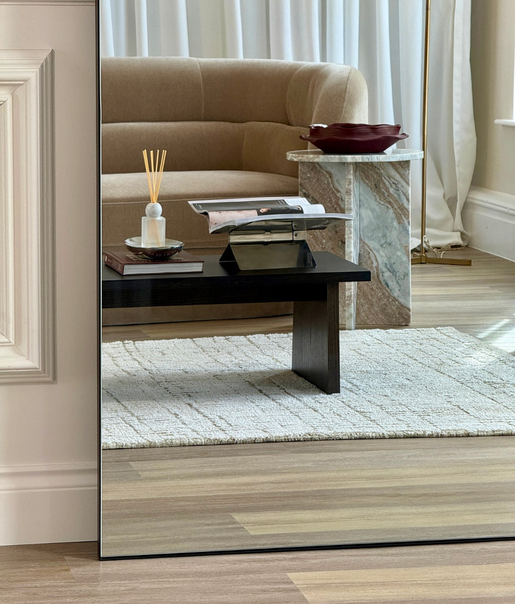 Reflection detail in arched full-length mirror with slim black frame showing living room sofa and coffee table