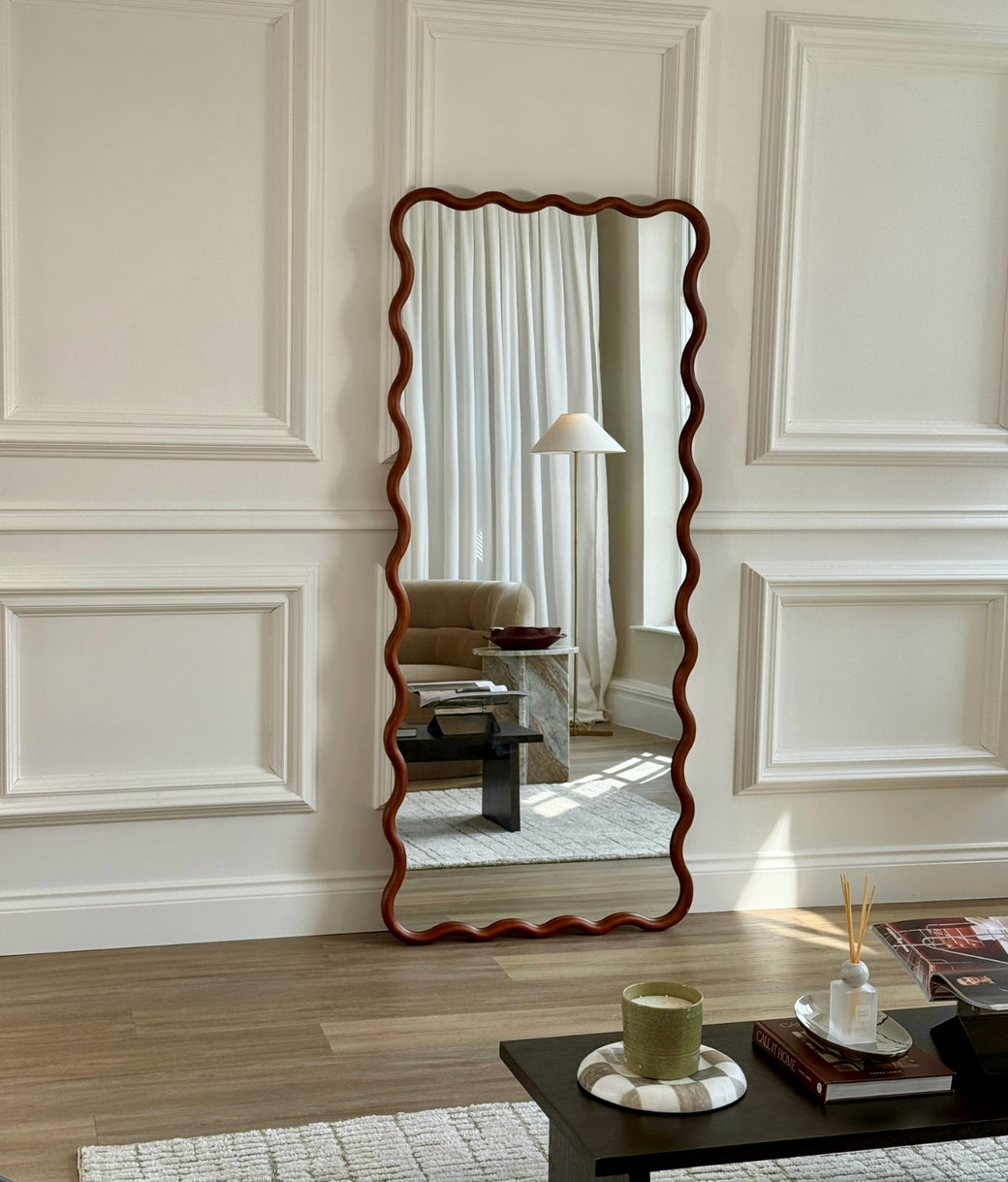 Full-length wavy frame mirror in walnut wood leaning against white panelled wall in living room