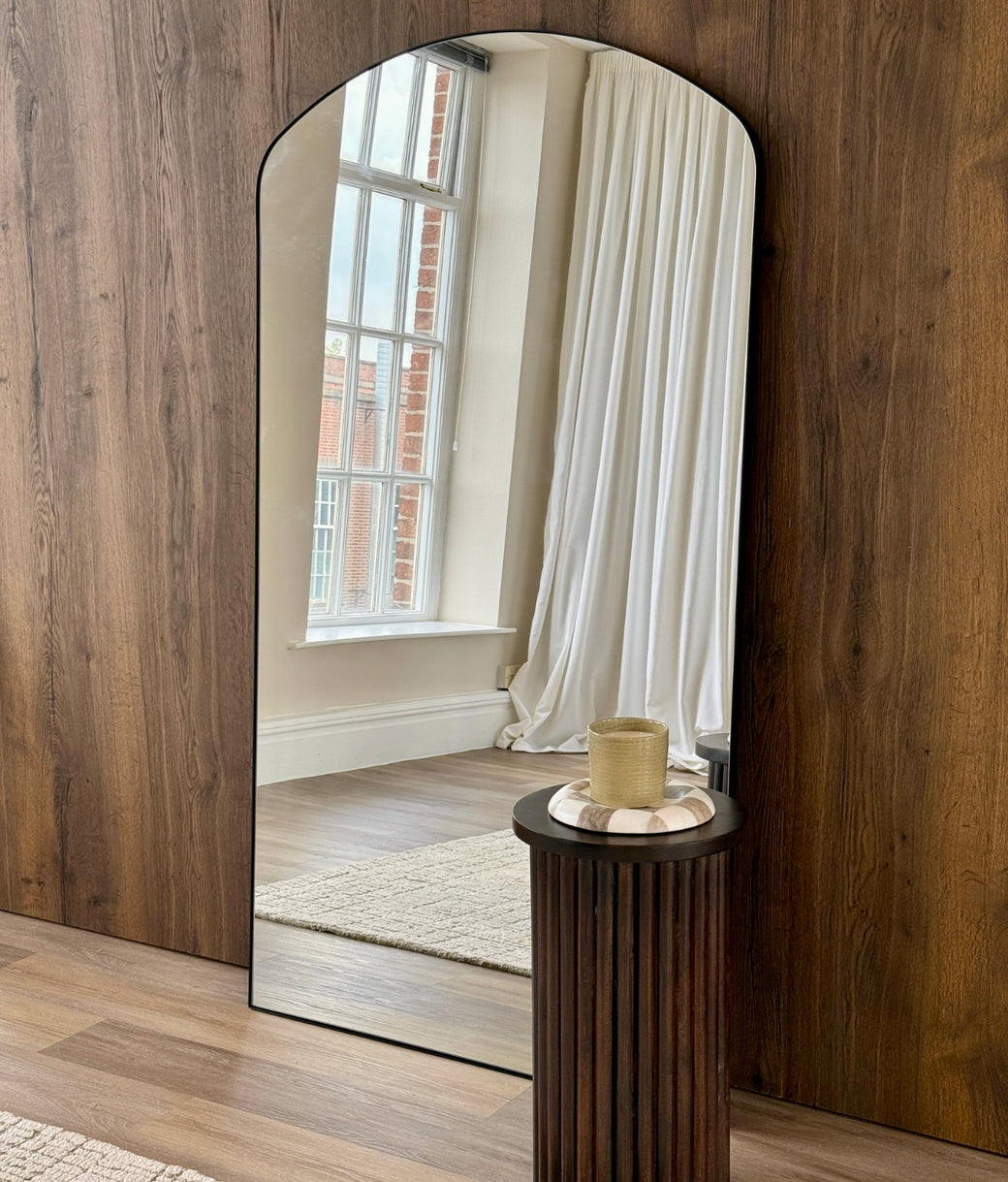 Full-length arched mirror with slim black frame against dark wood-panelled wall with reeded side table