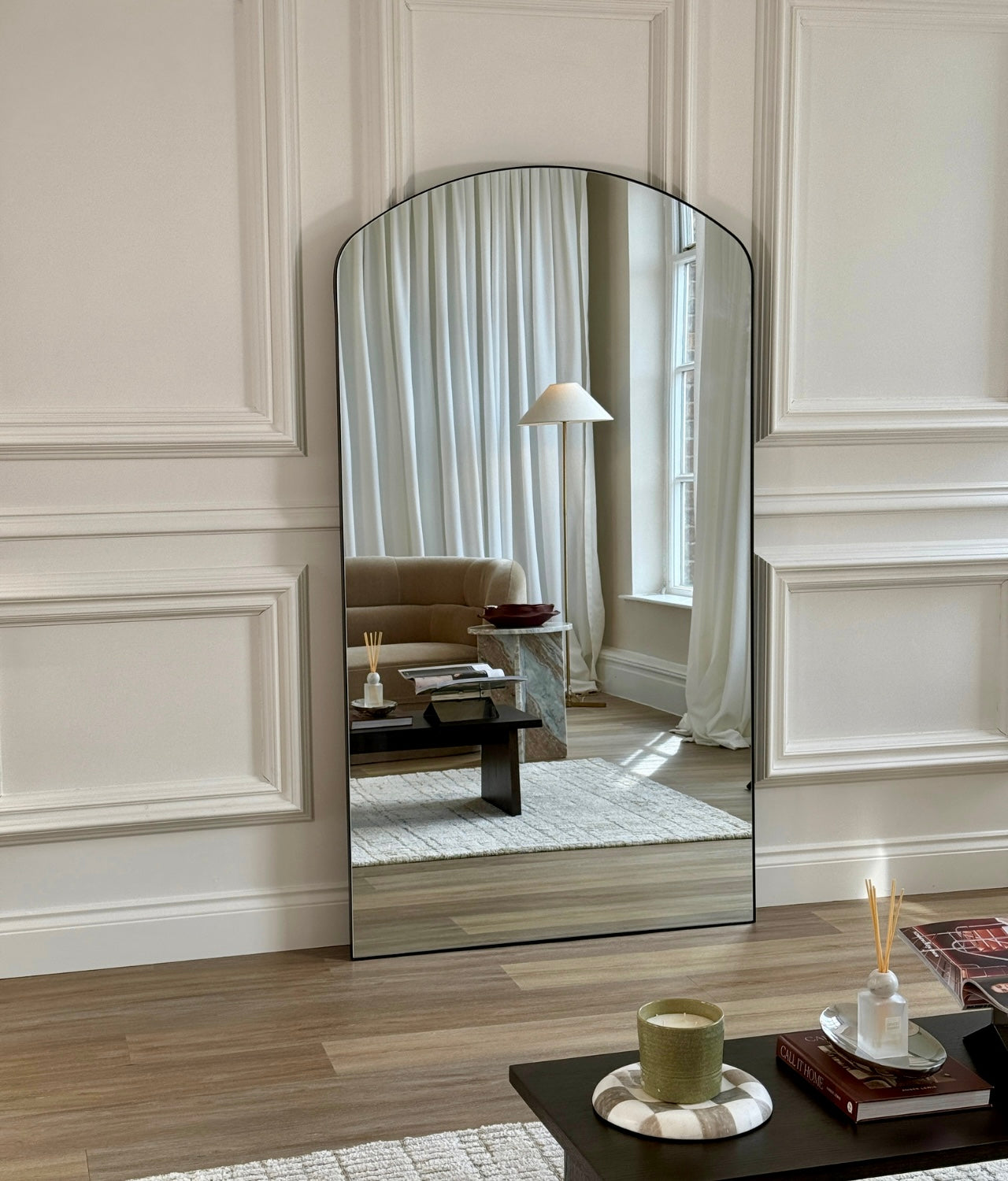 Arched full-length black frame mirror against white panelled wall reflecting floor lamp and living room
