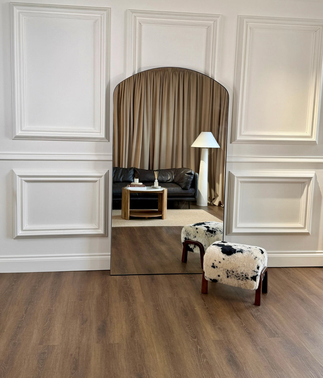 Wide view of full-length arched black frame mirror leaning against white panelled wall in styled living room