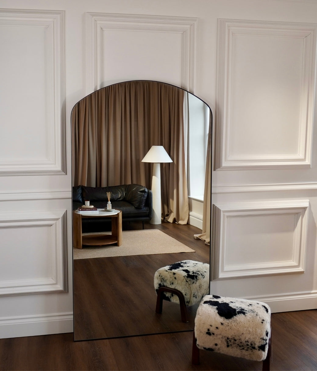 Wide view of full-length arched black frame mirror leaning against white panelled wall in styled living room