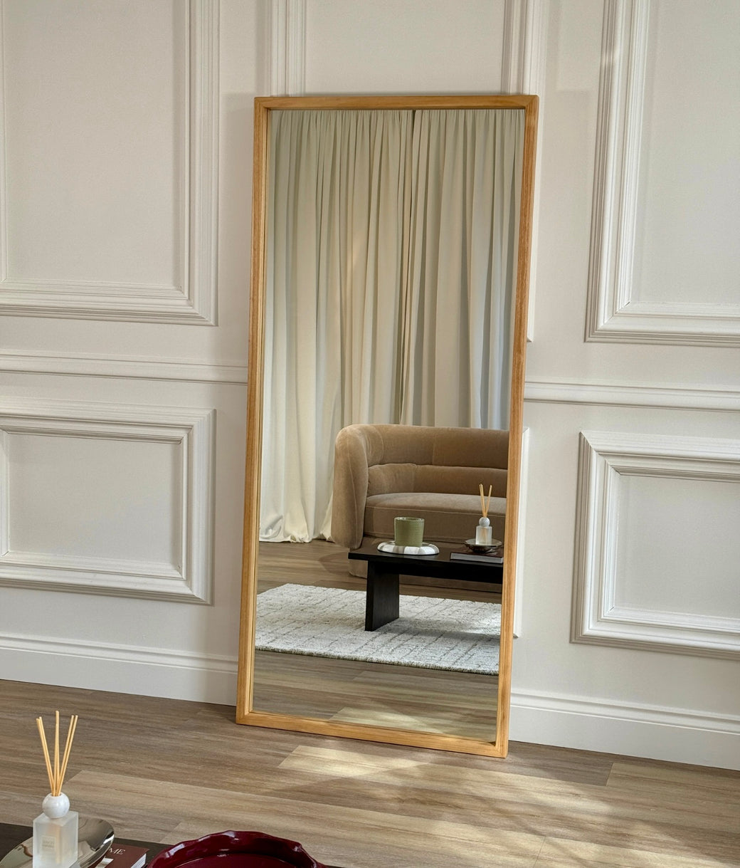 Full-length natural oak frame mirror leaning against white panelled wall with coffee table in foreground