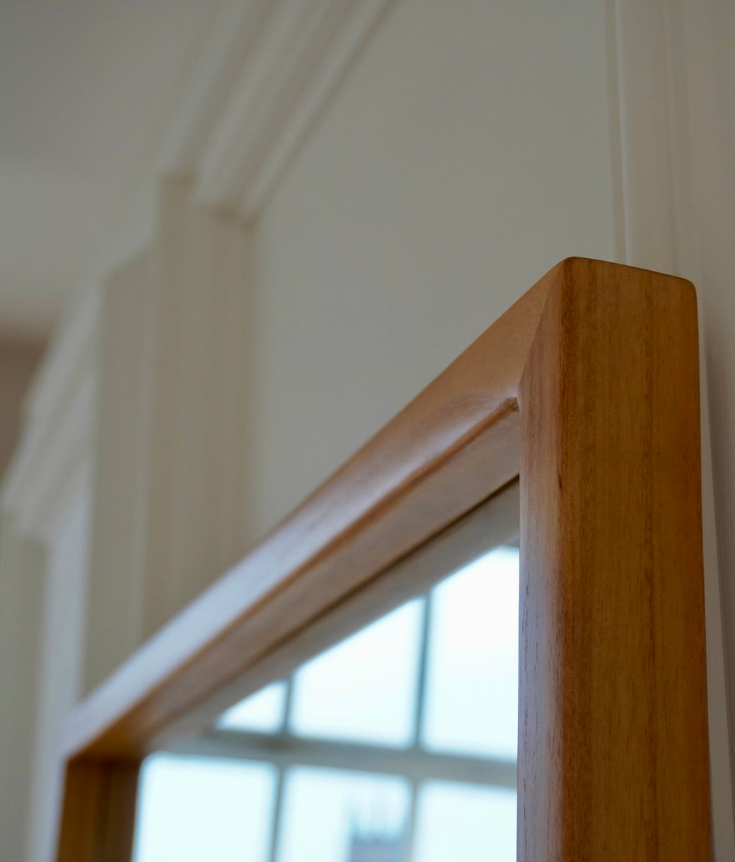Top corner detail of full-length mirror with natural oak wood frame against white panelled wall
