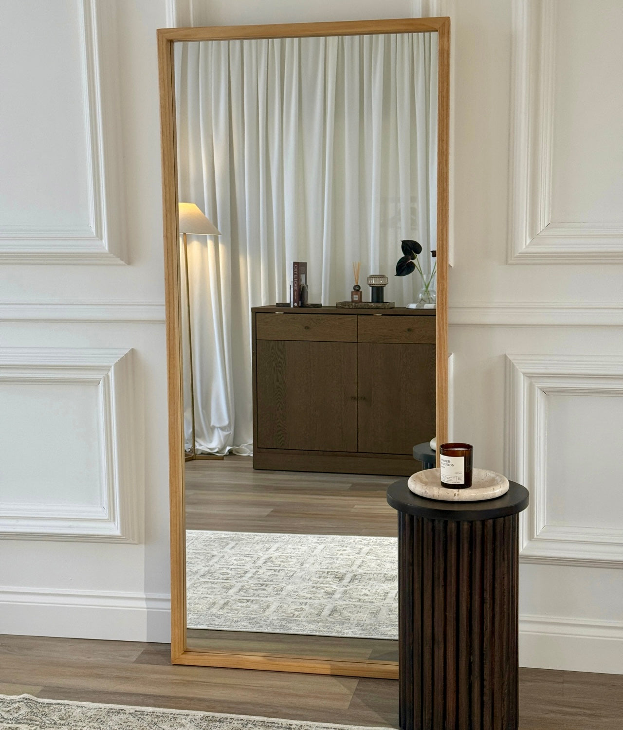 Full-length leaner mirror with natural oak frame leaning against white panelled wall in modern living room