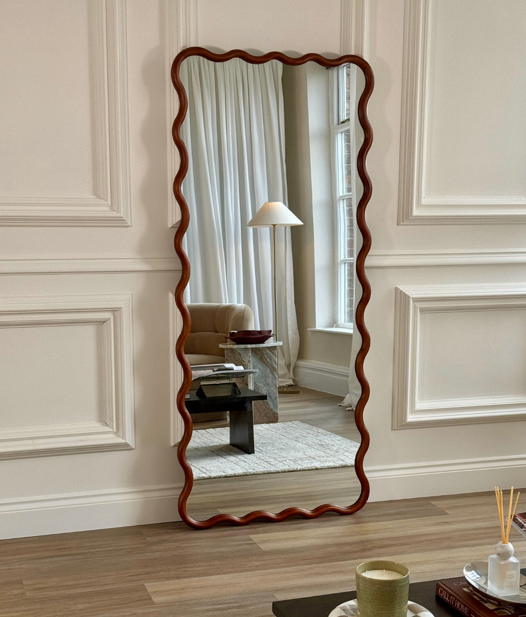 Full-length wavy wood frame mirror in walnut leaning against panelled wall with floor lamp in reflection