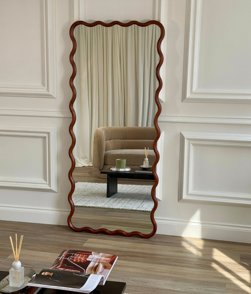 Full-length wavy walnut frame mirror standing against white panelled wall in styled living room
