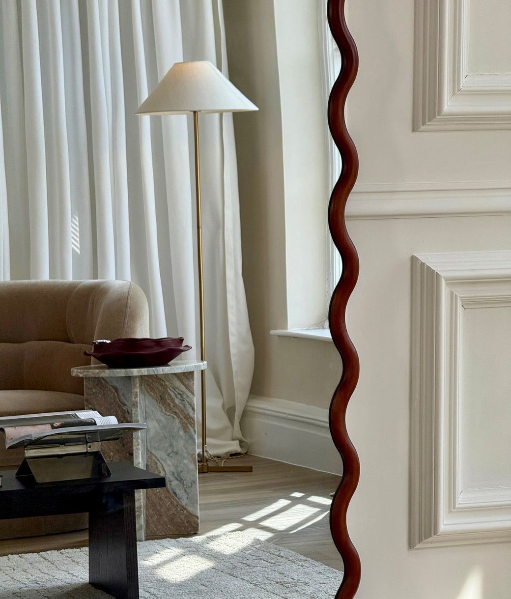 Side detail of wavy walnut wood frame mirror leaning against white panelled wall
